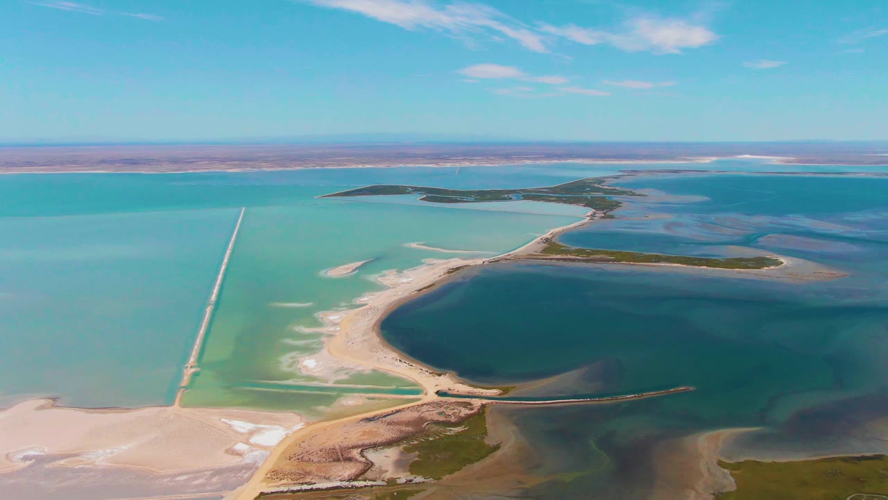 Drone view over Guerrero Negro with green-blue water sections, a central dark basin, pale sandbars, long dikes, distant low islands, and no visible people.