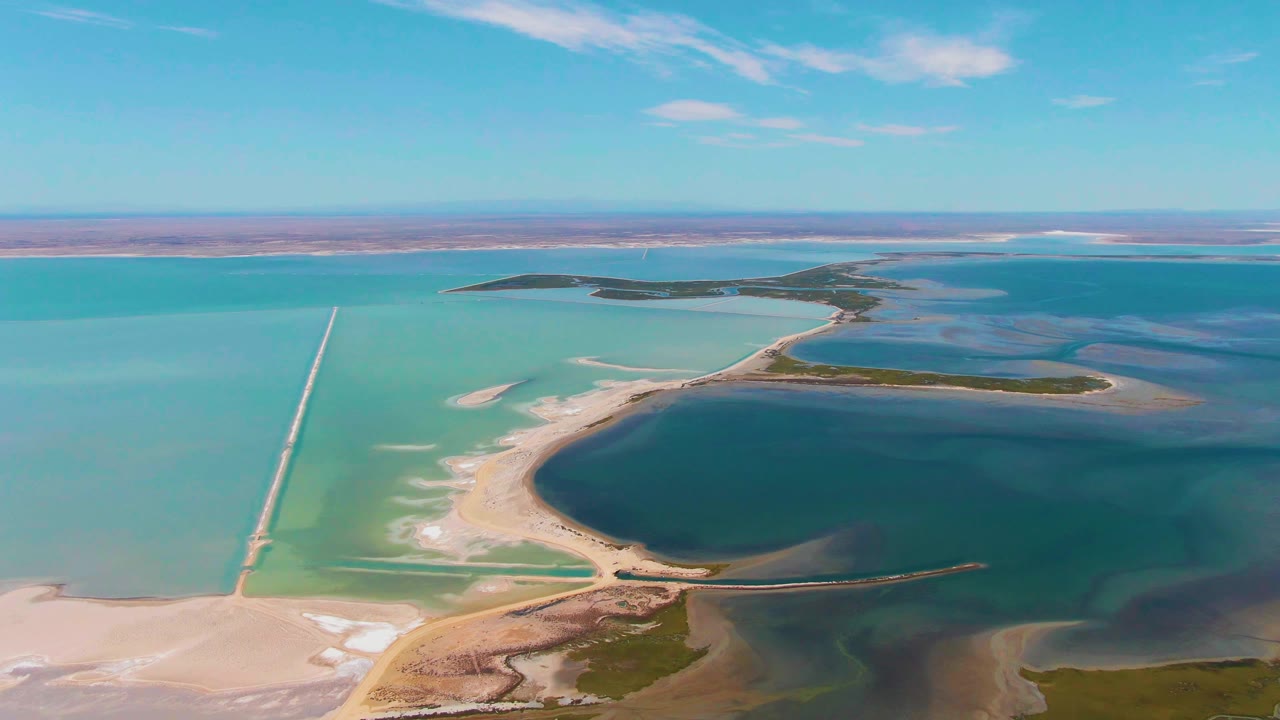 Wide aerial over Guerrero Negro salt flats with a straight pale causeway at left, curved sandy shoreline around dark water, turquoise basins, distant dry flats, and no visible people.
