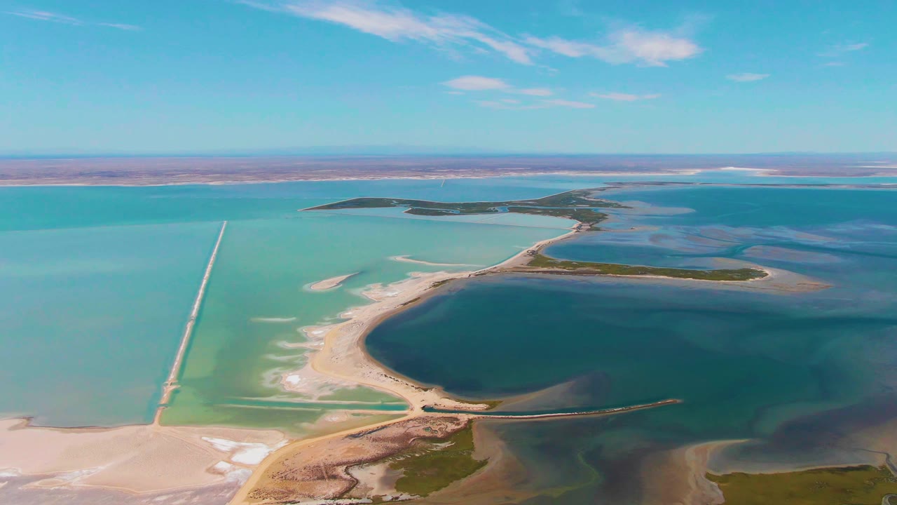 Aerial frame from Guerrero Negro showing blue-green basins divided by dikes, a pale causeway running through the left water, a dark central lagoon, and no visible people.