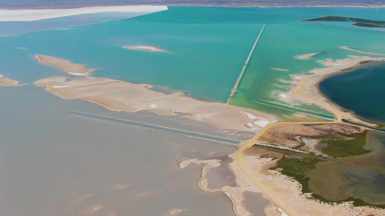 Aerial view in Guerrero Negro, Baja California Sur, with teal water across the top, gray-green shallows below, a curved shoreline at right, the long structure near center, and no visible people.