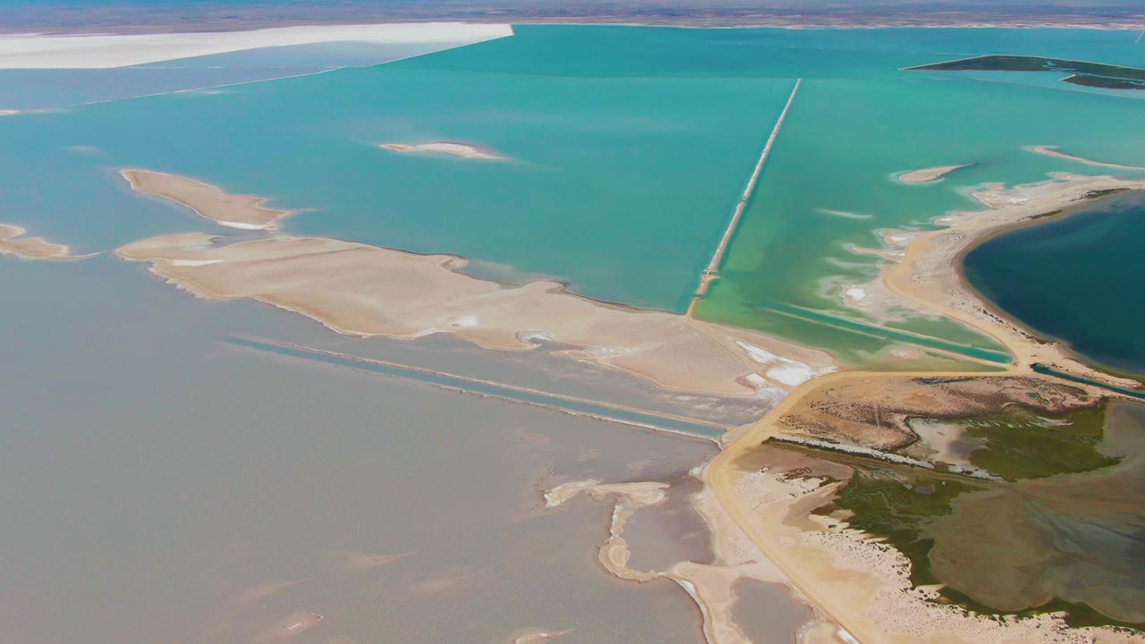 Guerrero Negro aerial frame showing sandy islands at left, a long straight structure through the turquoise shallows, darker water at right, green shoreline patches below, and no visible people.