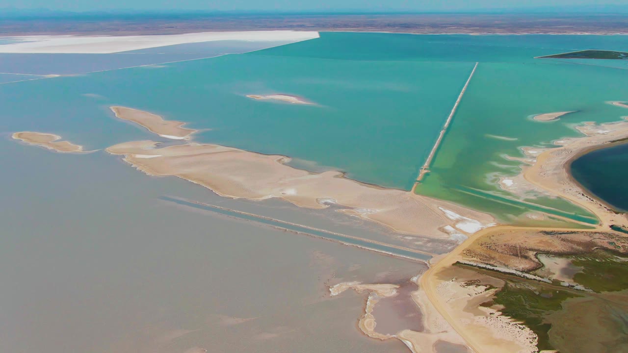 Aerial frame over Guerrero Negro, Baja California Sur showing turquoise water, pale sandy salt flats, white salt patches, a straight causeway-like line, and no visible people.