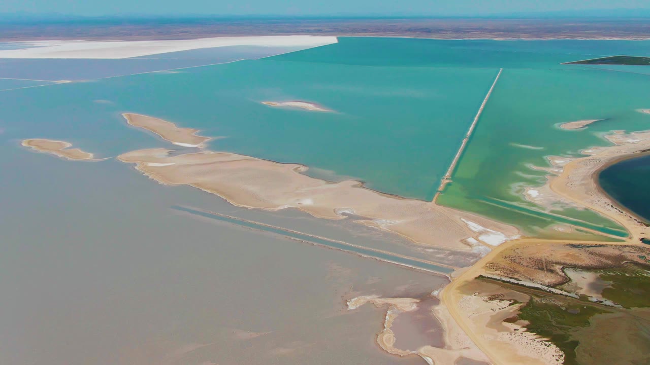 Second aerial frame from Guerrero Negro with pale sand bars surrounded by turquoise and gray water, a long narrow structure crossing the water, and no visible people.