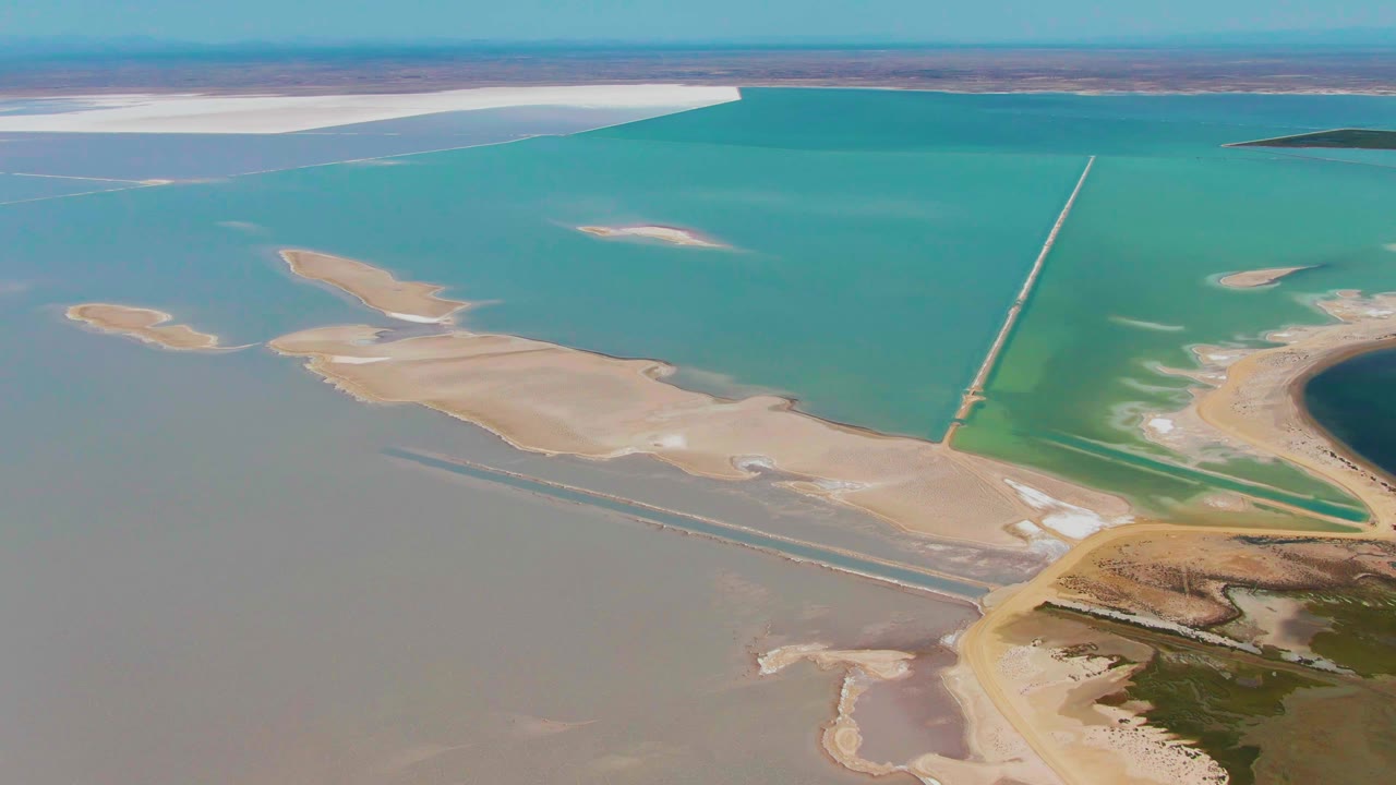 Wide aerial view in Guerrero Negro showing submerged salt-flat edges, turquoise water, a long causeway or pipeline-like feature, distant shoreline, and no visible people.