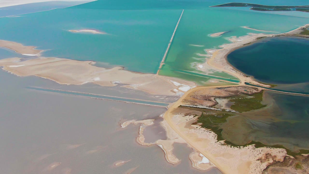 Opening aerial frame over Guerrero Negro, Baja California Sur, showing turquoise saltwater, pale sandbars, narrow causeways, green wetland patches, a dark circular pond, and no visible people.
