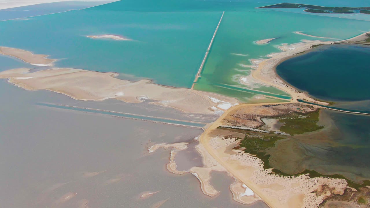 Second aerial frame over Guerrero Negro wetlands with bright turquoise water above pale sand flats, a causeway cutting through the lagoon, the dark pond at right, and no visible people.