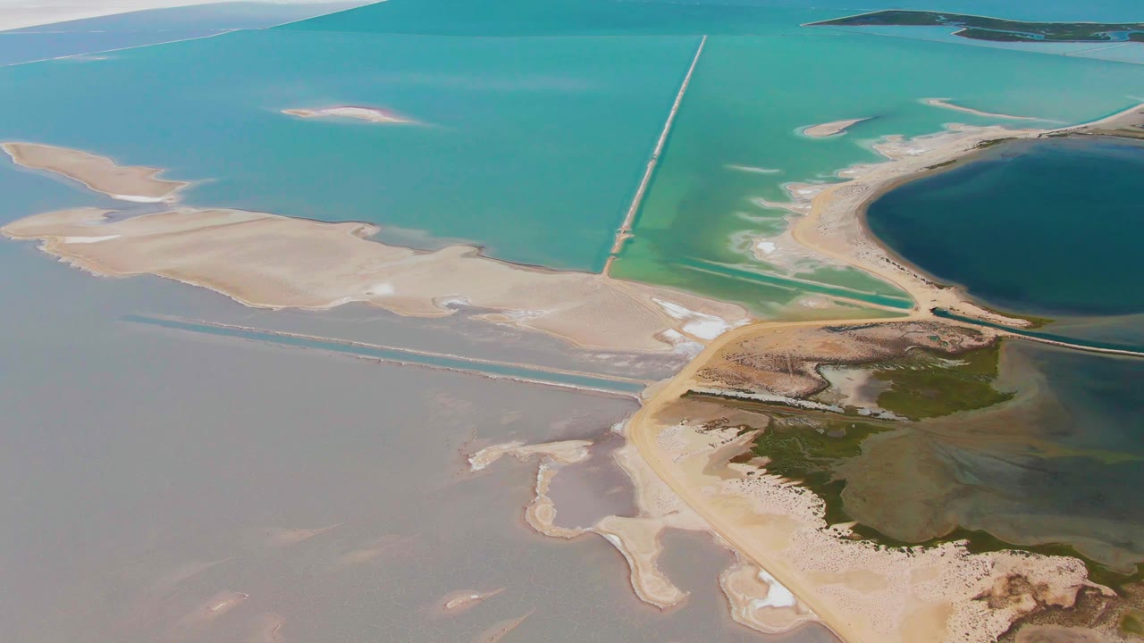 Third frame from the Guerrero Negro aerial clip showing the central sandbar area, straight causeway lines through blue-green water, green wetland below, the dark circular pond at right, and no visible people.