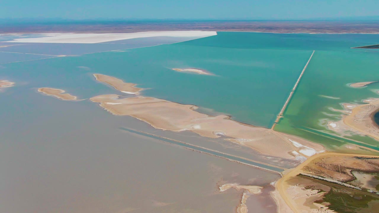 Opening aerial frame over Guerrero Negro evaporation ponds with turquoise water, beige sandy islands, white salt deposits near the horizon, and no visible people.