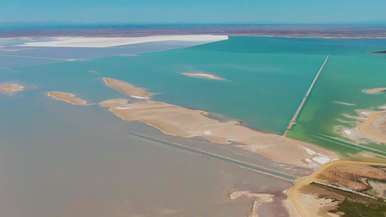 Aerial frame at 0:02 in Guerrero Negro with turquoise evaporation ponds filling the frame, pale salt deposits along the far side, sandy islands in the center, and no visible people.