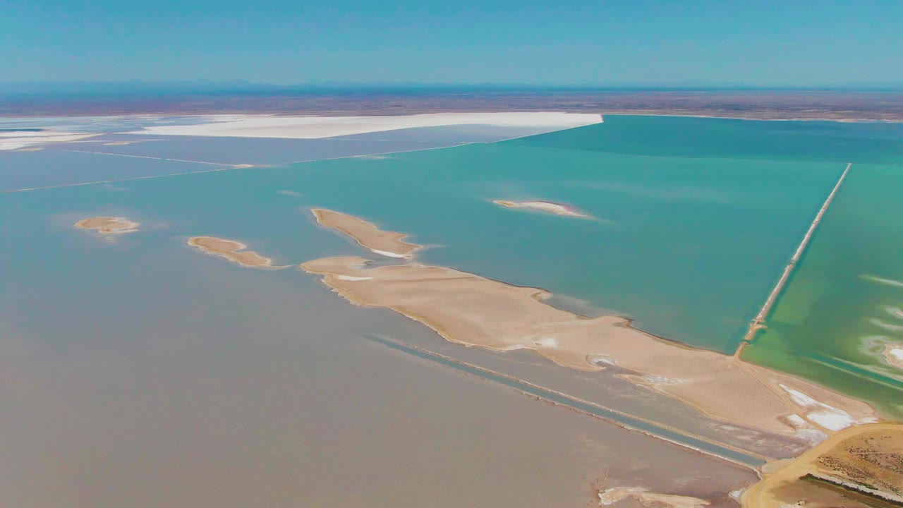 High aerial frame of Guerrero Negro evaporation ponds with a broad turquoise water body, pale white salt flats near the horizon, brown water below, small sand islands, and no visible people.