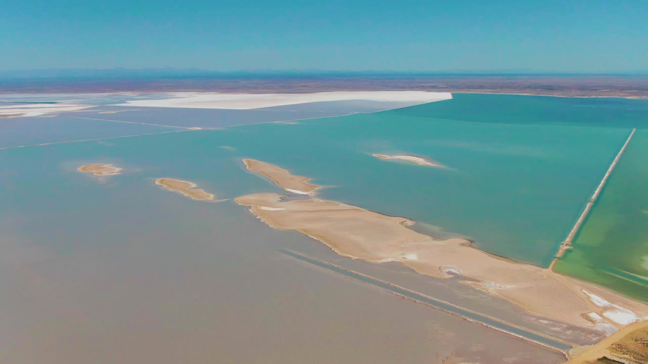 Wide aerial frame over Guerrero Negro showing turquoise water, tan land patches in the foreground, white salt flats near the horizon, a straight causeway at right, and no visible people.