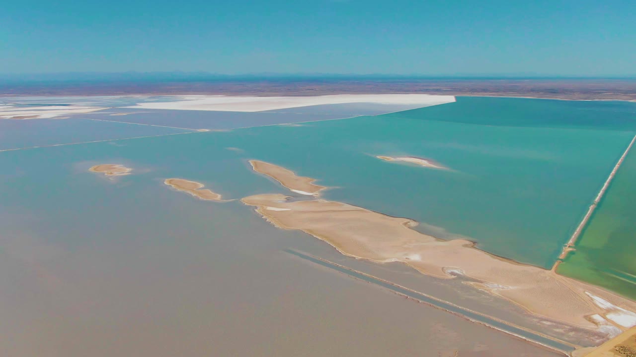 Guerrero Negro aerial frame with white salt flats stretching along the far shoreline, turquoise water below them, tan land patches lower center, the causeway at right, and no visible people.