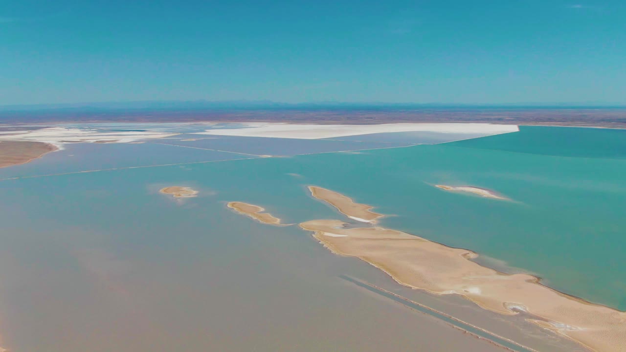 Opening aerial frame over Guerrero Negro, Baja California Sur, with turquoise water surrounding pale sandy islands, white salt-flat edges on the horizon, and no visible people.