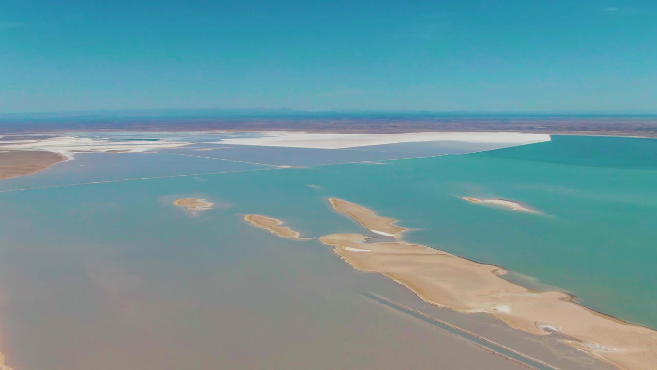 Aerial frame in Guerrero Negro showing gray-green shallows at left, brighter turquoise water at right, sandy island shapes through the center, distant white flats, and no visible people.