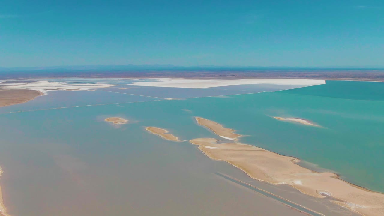 Third frame of the Guerrero Negro aerial view, with narrow pale sandbars stretching across turquoise water, a broad white shoreline band in the distance, and no visible people.