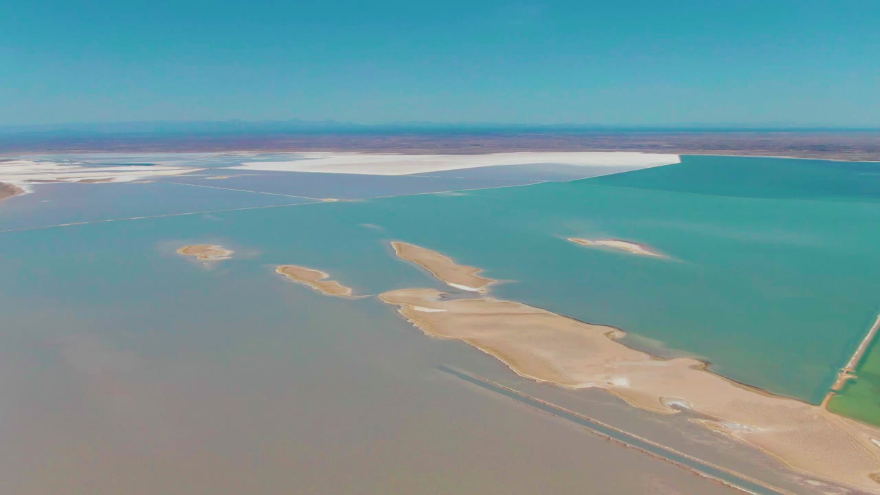 Opening aerial frame over Guerrero Negro salt flats, with turquoise water ponds, tan sandbars, white salt beds near the horizon, clear blue sky, and no visible people.