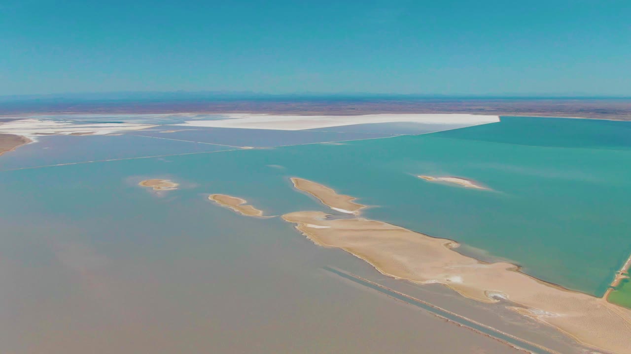 Aerial view over Guerrero Negro with tan landforms crossing turquoise water, white salt areas along the far shoreline, a straight horizon, and no visible people.