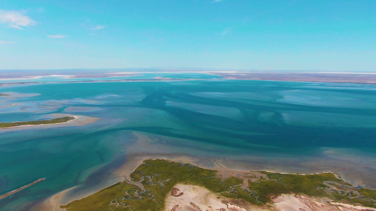 At three seconds, the aerial composition over Guerrero Negro keeps the shoreline low in frame, with broad shallow water, sandbar shapes, distant flats, and no visible people.