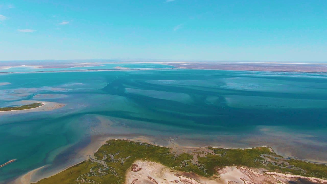 Wide aerial of coastal inlet water, sandy edges, vegetation, and a low horizon.