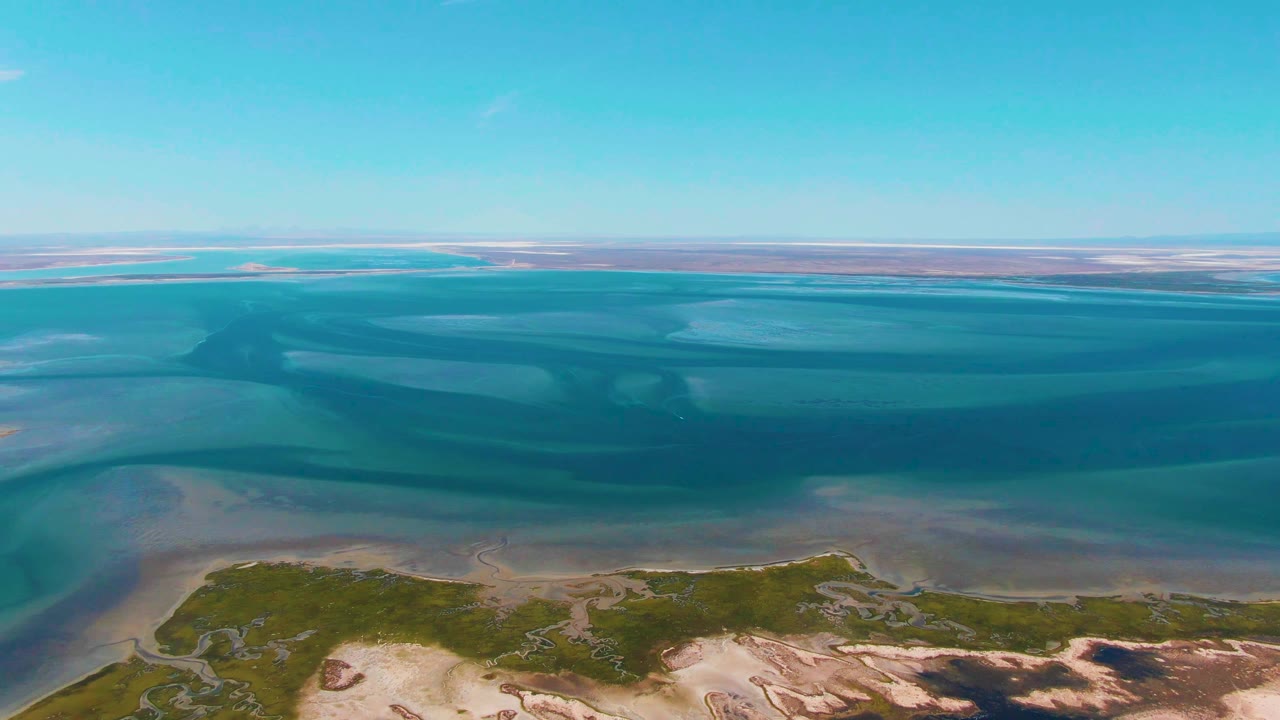 Opening aerial frame over Guerrero Negro coastal waters, with turquoise shallows, darker tidal channels, sandy shoreline, green vegetation, and no visible people.
