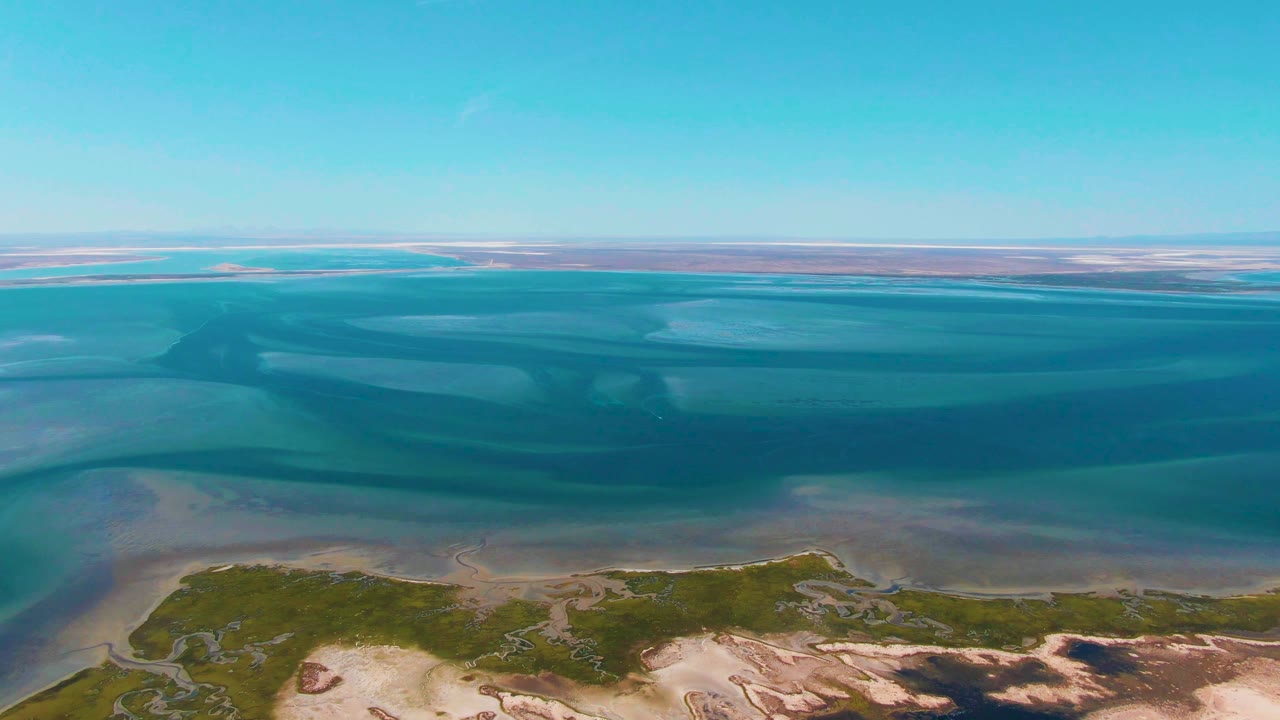 A second aerial moment over Guerrero Negro shows layered blue and turquoise water bands above a sandy vegetated shoreline, with no visible people.