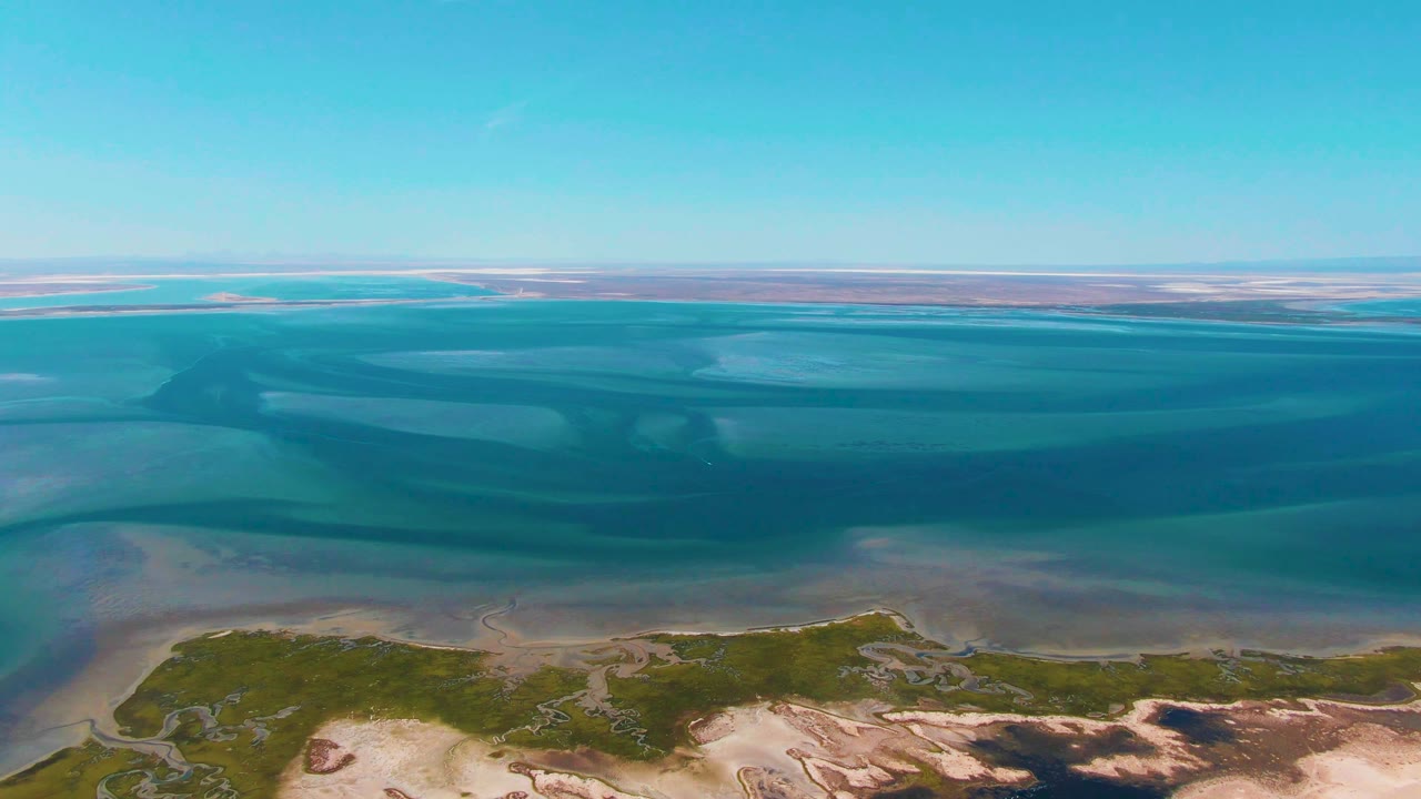 The aerial view continues over Guerrero Negro coastal shallows, with pale sandbars under clear turquoise water and a low shore at the bottom, with no visible people.