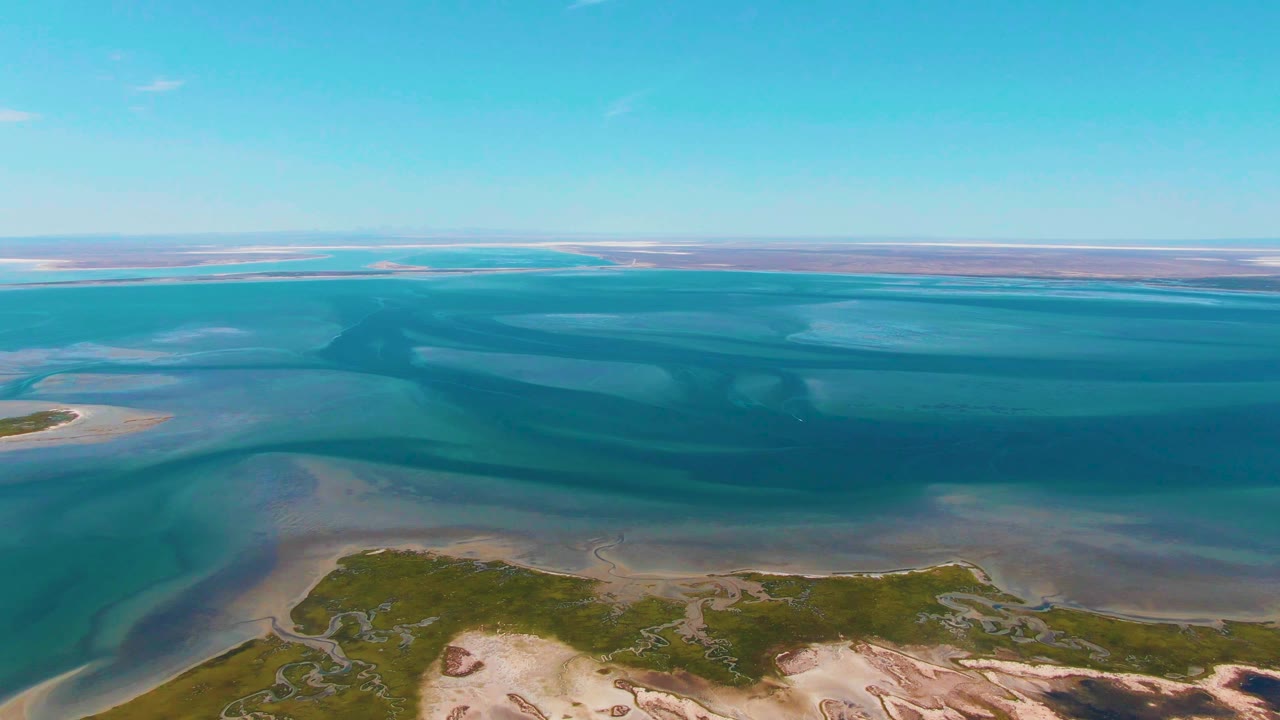 Aerial view over Guerrero Negro coastal wetlands with turquoise water bands, pale sandbars, green shoreline vegetation at the bottom edge, and no visible people.
