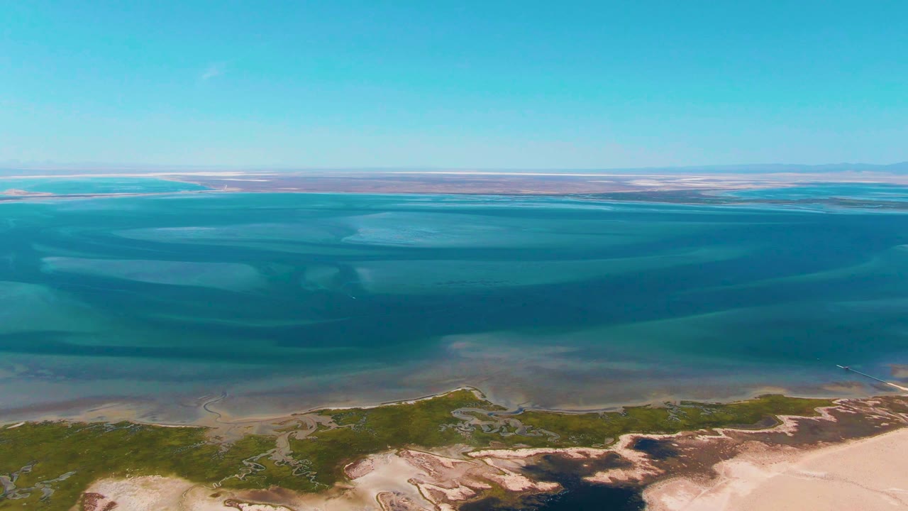 High aerial frame over Guerrero Negro, Baja California Sur shows layered blue-green lagoon water, pale sand, low shoreline vegetation, distant horizon, and no visible people.
