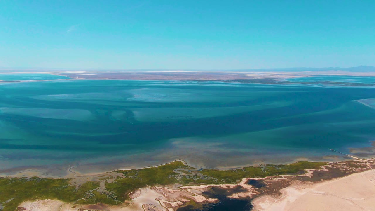 The Guerrero Negro aerial frame shows darker teal bands crossing the lagoon, pale sand at the lower right, scattered green coastal vegetation, and no visible people.