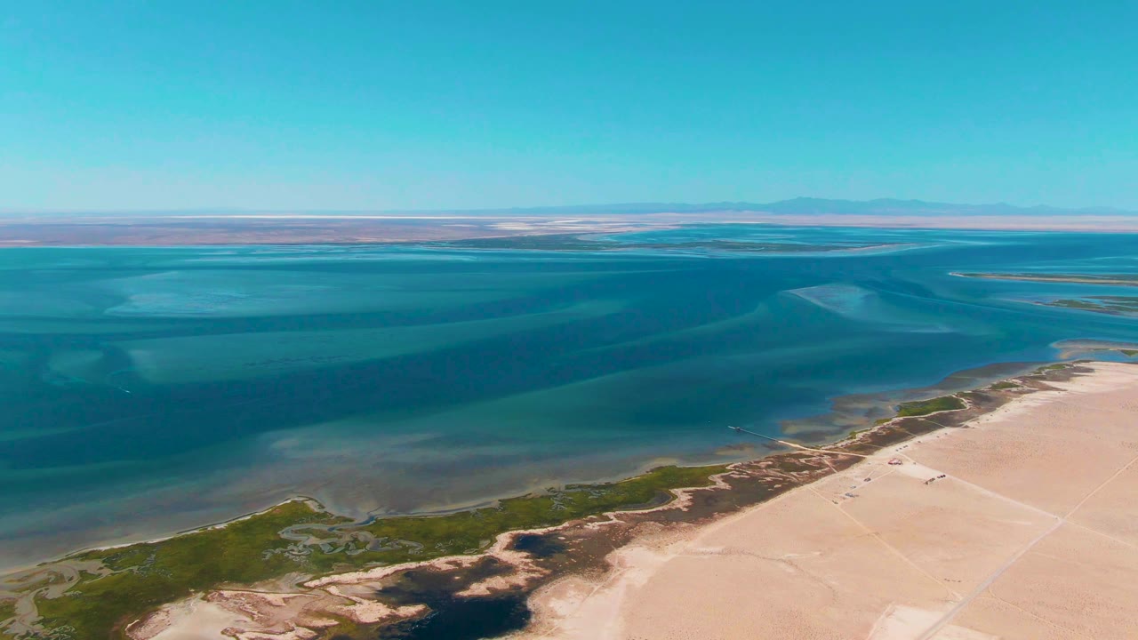 Opening aerial frame over Guerrero Negro, Baja California Sur, with blue green water, pale sandy shore, low vegetation along the edge, distant mountains, and no visible people.