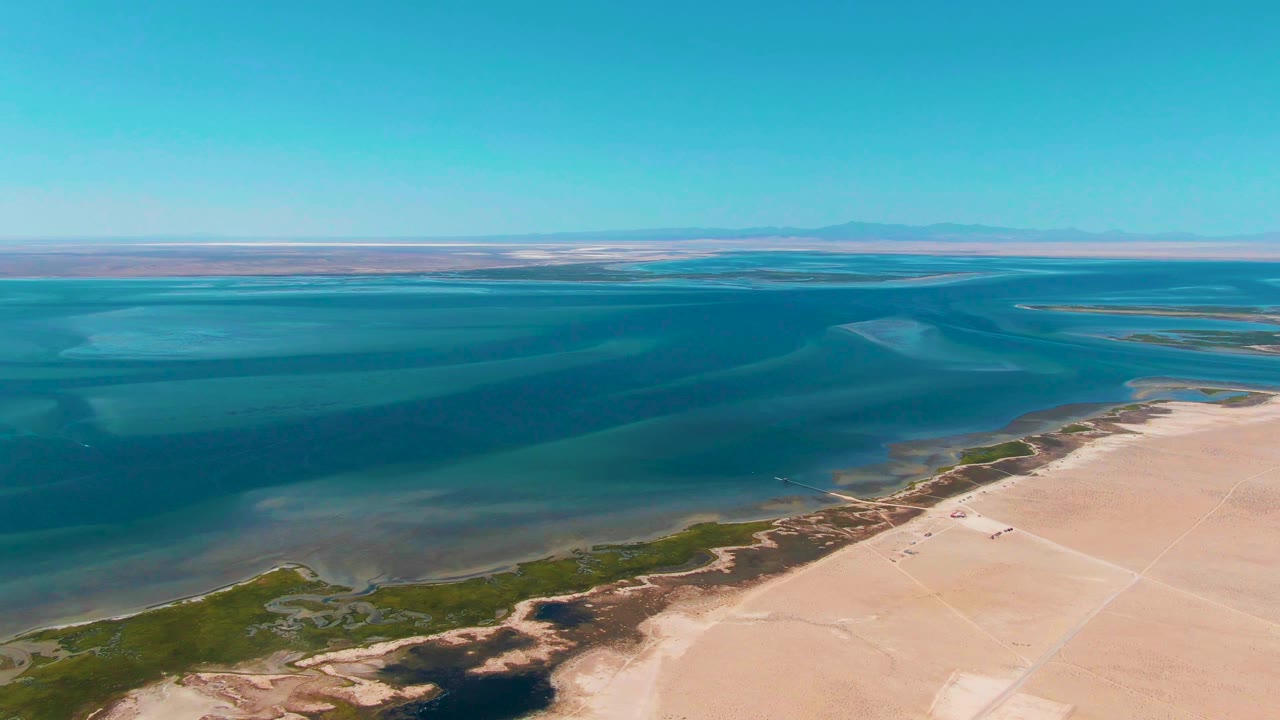 Mid-clip aerial frame over Guerrero Negro, Baja California Sur, with turquoise water, a sandy shoreline, sparse green edge vegetation, a tiny vehicle on the sand, and no visible people.