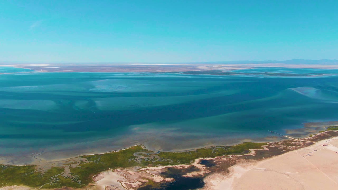Aerial frame over Guerrero Negro, Baja California Sur, showing turquoise coastal water, pale sandy shore, green vegetation patches, a clear blue sky, and no visible people.