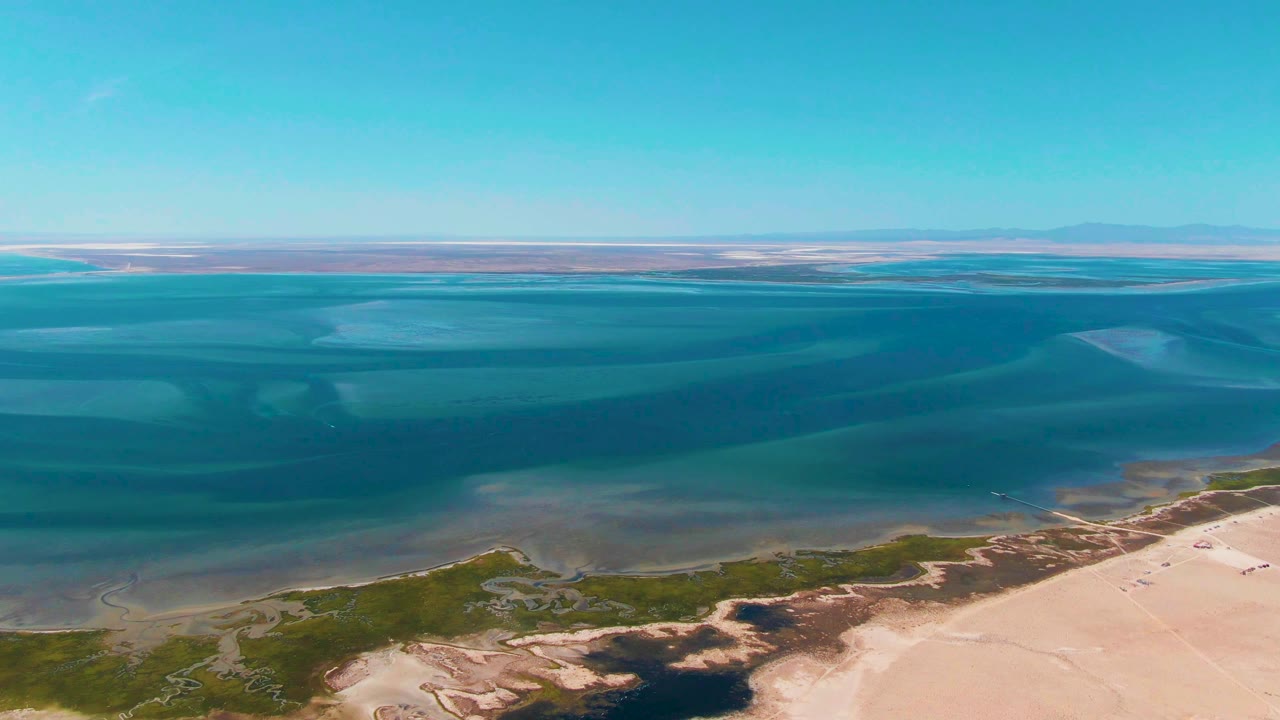 Aerial view in Guerrero Negro, Baja California Sur, with layered blue water across the middle of frame, pale sand at the lower right, green shoreline patches, distant land, and no visible people.