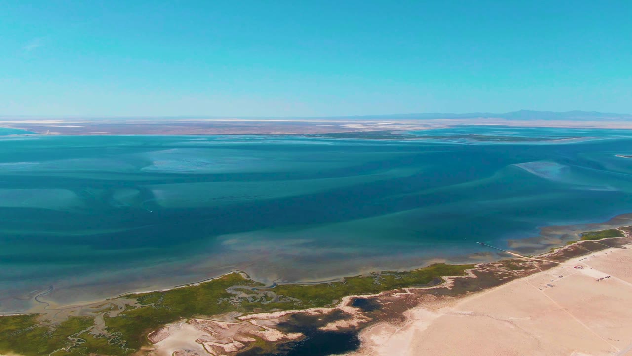 Aerial Guerrero Negro shoreline frame showing darker blue water offshore, lighter shallows near the sand, green patches along the beach edge, clear sky, and no visible people.