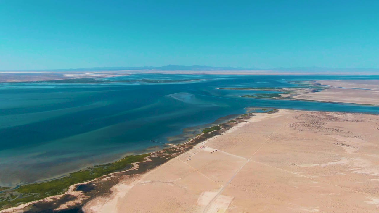 Aerial view over Guerrero Negro, Baja California Sur, showing pale sandy shore, blue ocean water, green shoreline vegetation, distant mountains, and no visible people.