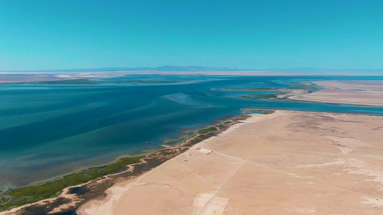 High aerial frame over Guerrero Negro with the shoreline running diagonally between pale sand, dark blue water, green edge vegetation, distant mountains, and no visible people.