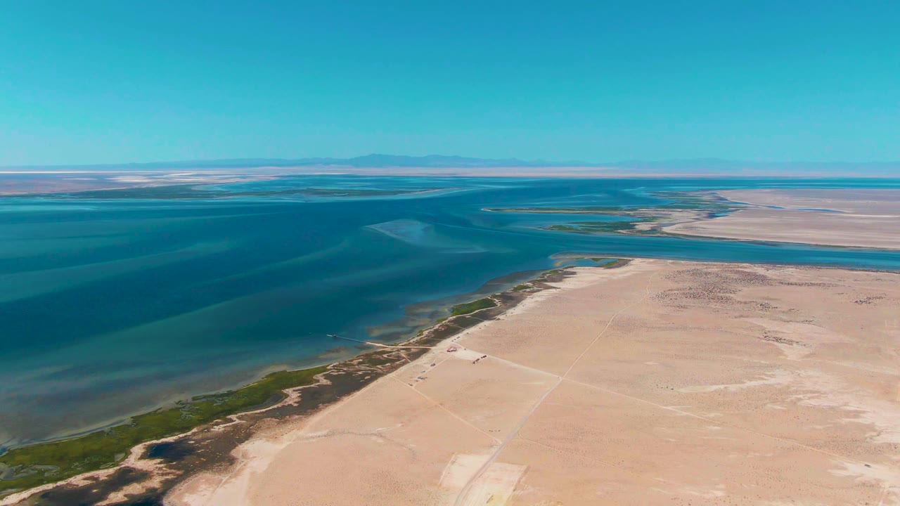 Aerial view in Guerrero Negro showing coastal desert sand in the foreground, blue-green ocean bands in the midground, distant mountains on the horizon, and no visible people.