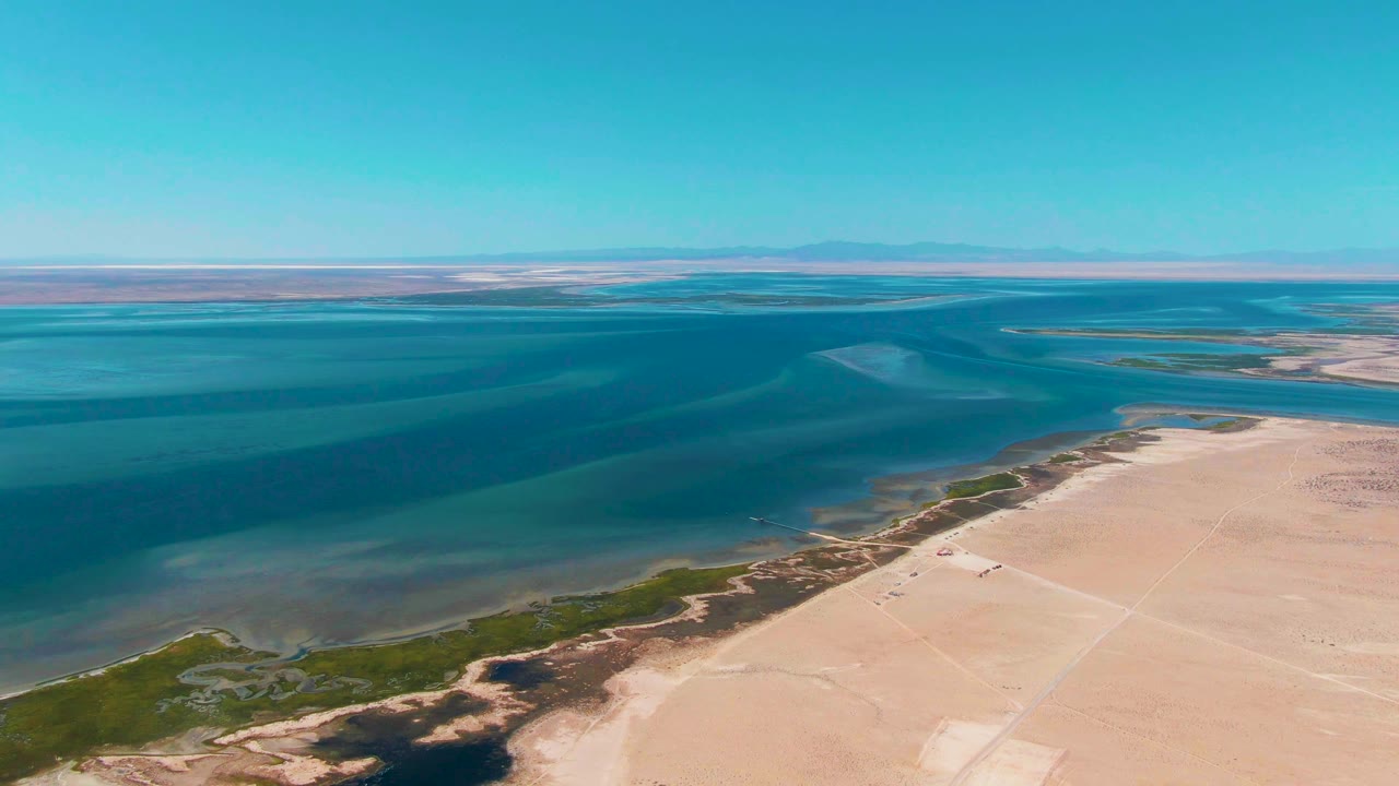Aerial view over Guerrero Negro coastal water with pale sandy shoreline, green edge vegetation, distant horizon, clear sky, and no visible people at the start of the clip.