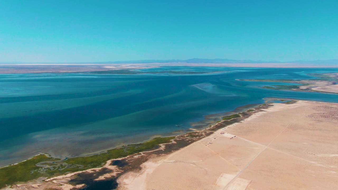 Aerial frame over Guerrero Negro showing darker blue water bands, a pale sandy shore on the right, thin green vegetation along the waterline, clear sky, and no visible people.