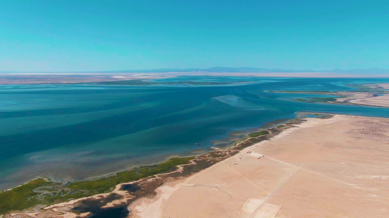 Wide aerial view in Guerrero Negro with teal and dark blue coastal water, sandy desert land on the right, a narrow green shoreline strip, distant mountains, and no visible people.