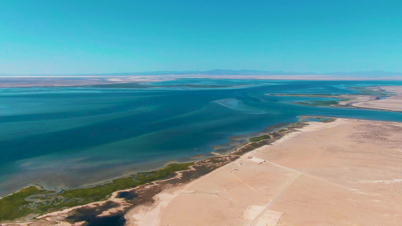 Aerial view across Guerrero Negro shoreline where shallow green water shifts into deeper blue, with pale sand flats, low vegetation near the edge, clear sky, and no visible people.