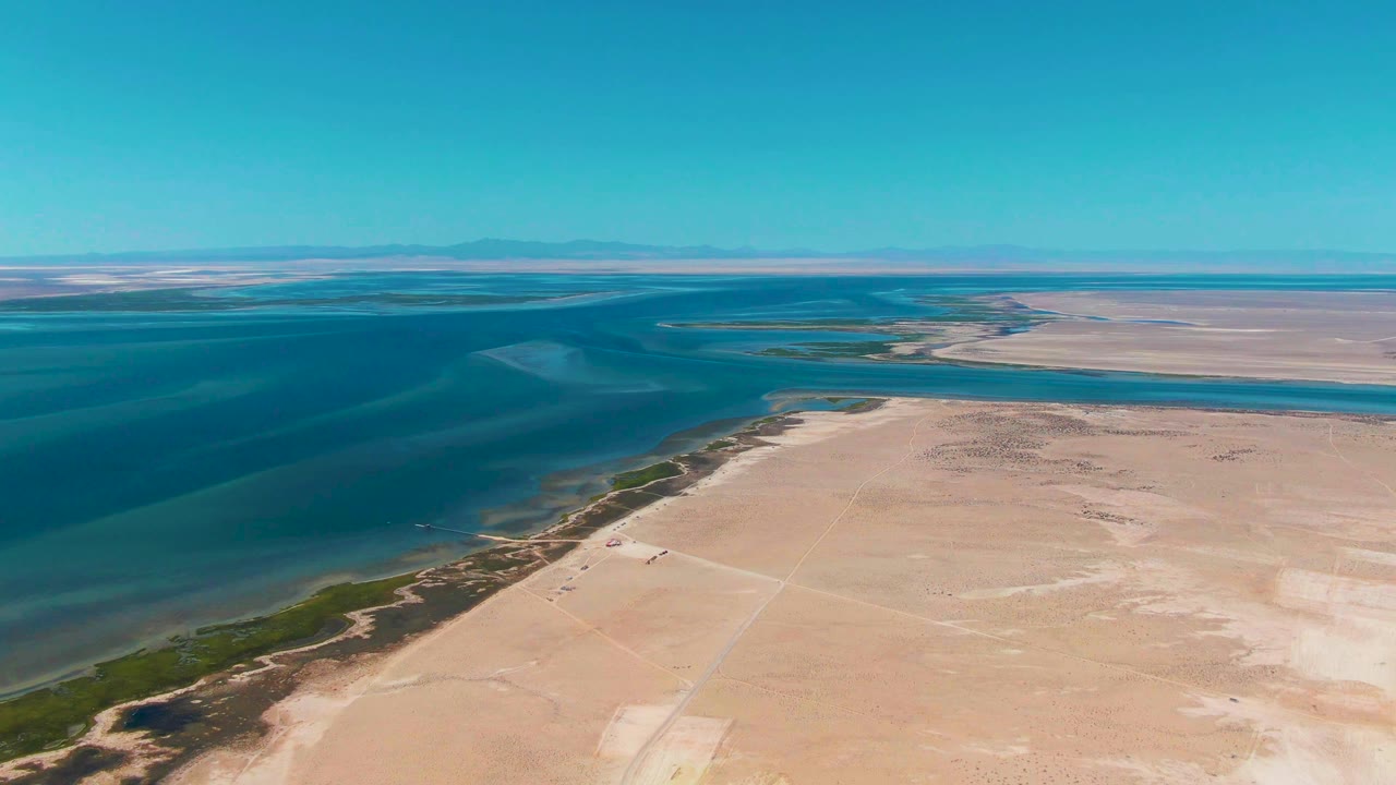 Aerial frame over Guerrero Negro, Baja California Sur, showing turquoise coastal water on the left, pale sandy desert in the foreground, low shoreline vegetation, a flat distant horizon, and no visible people.