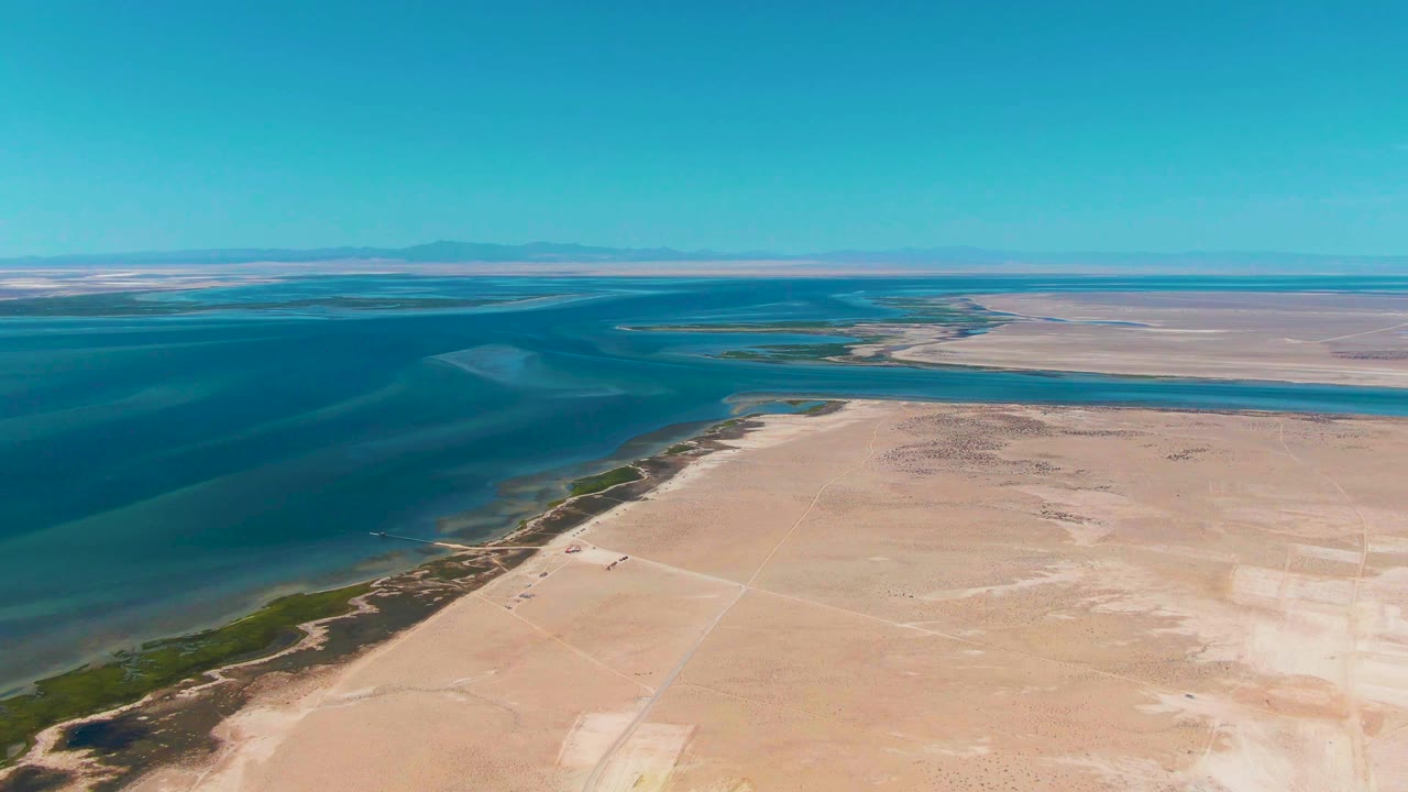 High aerial frame over Guerrero Negro, Baja California Sur, showing calm blue ocean on the left, pale sand flats on the right, a curving vegetated shoreline, hazy distant horizon, and no visible people.