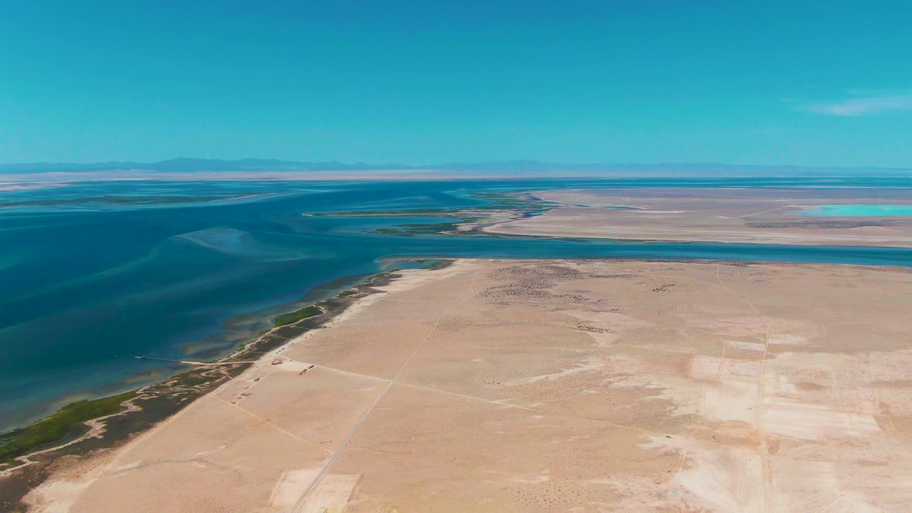 Opening aerial frame over Guerrero Negro, Baja California Sur, with blue water along a pale sandy shore, dry flat land in front, low horizon in back, and no visible people.