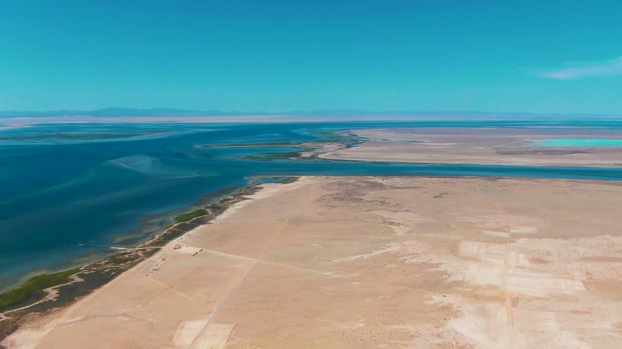 Aerial view one second into the Guerrero Negro clip, with the blue waterline curving along the left side, pale sand spreading across the foreground, distant land on the horizon, and no visible people.
