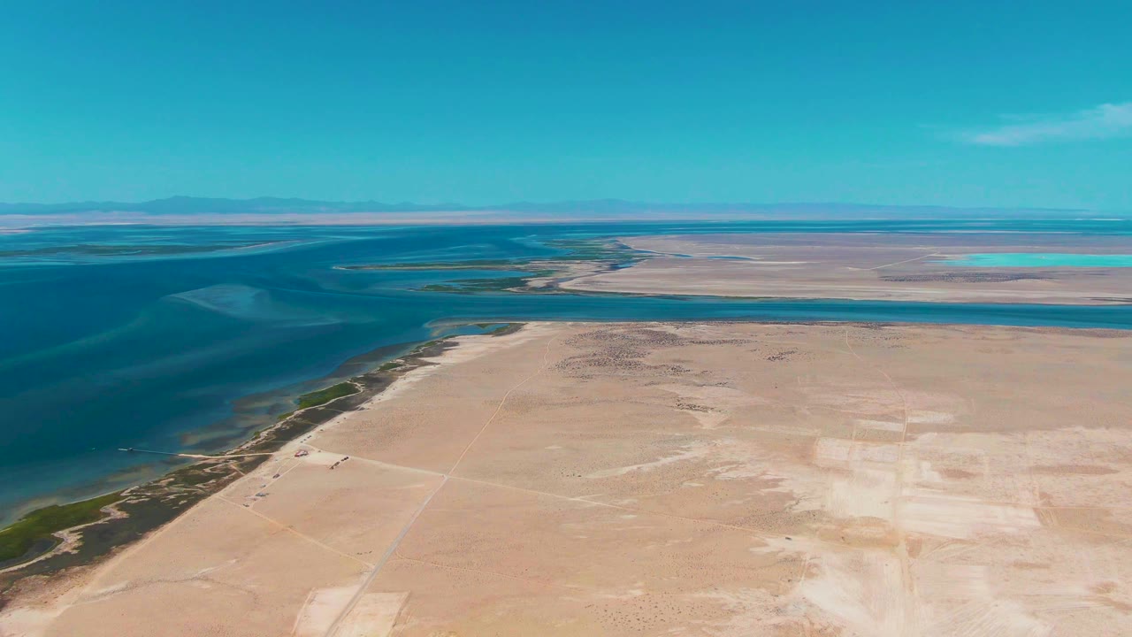 Two seconds in, the aerial frame shows dark blue coastal water on the left, a narrow green edge at the shore, pale dry flats across the lower frame, distant flat land, and no visible people.