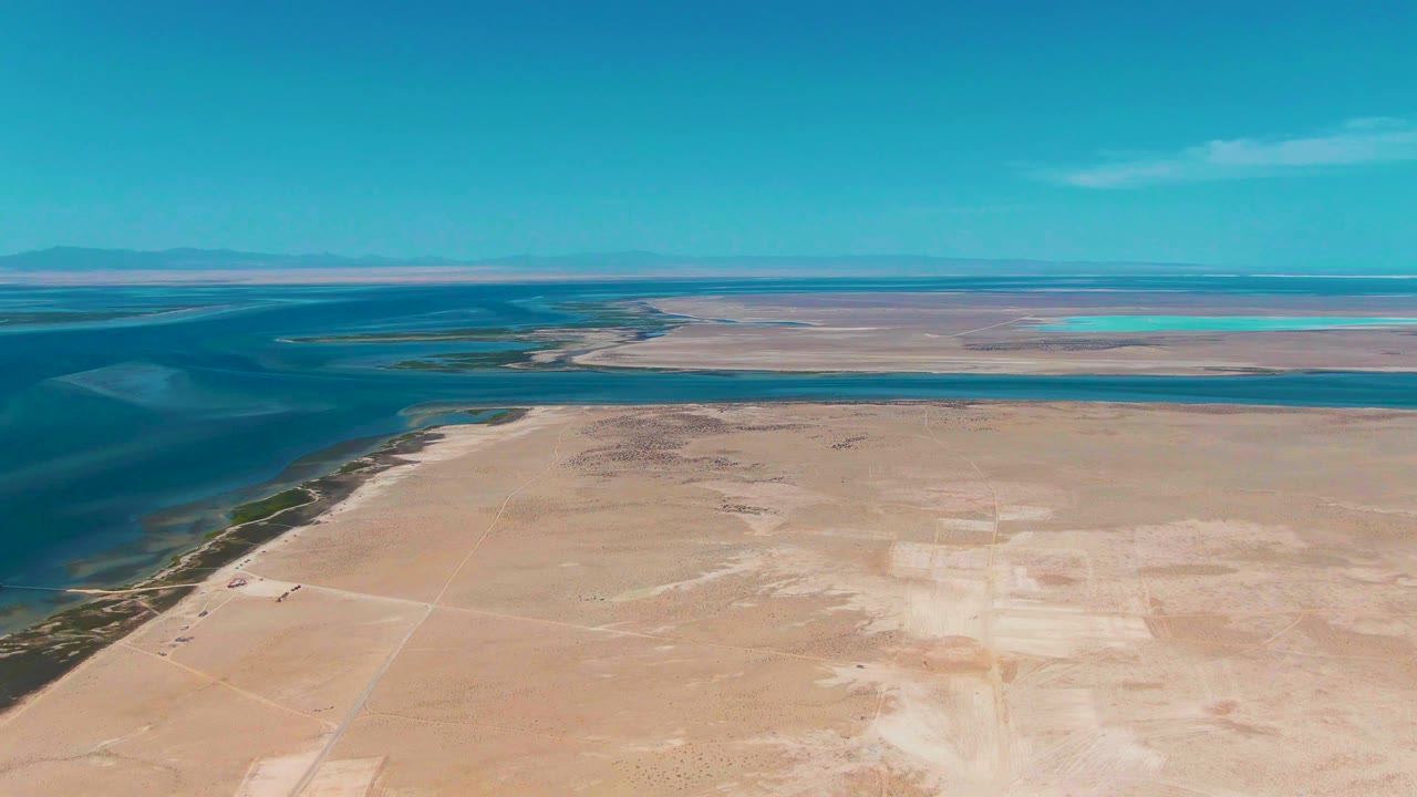 Early frame over Guerrero Negro showing darker lagoon water along the left edge, pale arid flats across the foreground, low mountains on the horizon, and no visible people.
