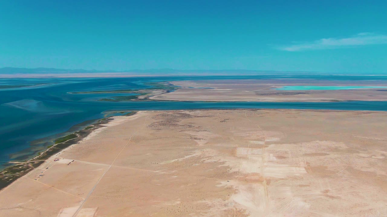Aerial frame in Guerrero Negro with the lagoon curving along the left, sandy shoreline below, turquoise water bands in the distance, and no visible people.