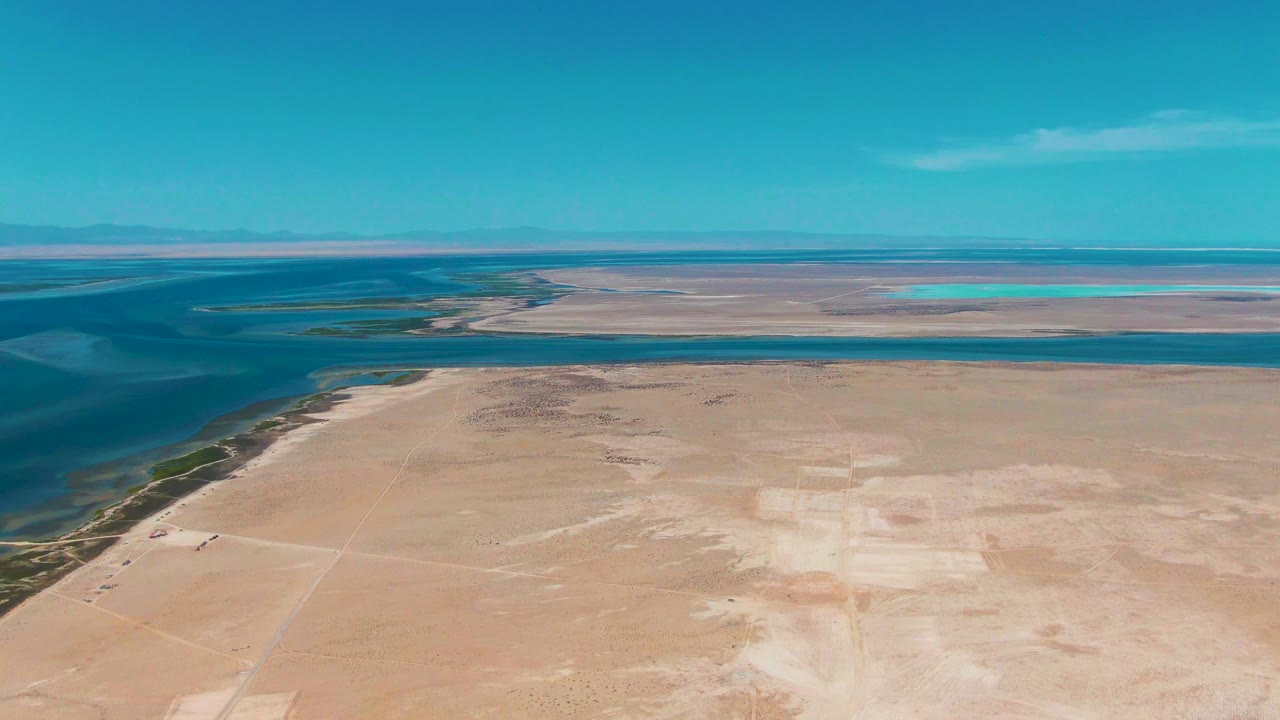 Mid-clip aerial view of Guerrero Negro with calm blue water bordering the left side of a broad sandy plain, distant flat wetlands, and no visible people.
