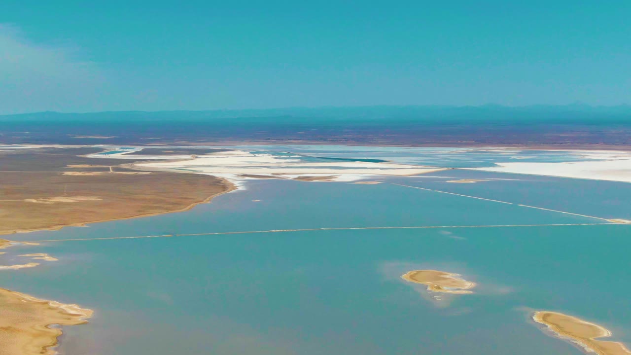 Aerial view of turquoise water, white salt flats, tan shoreline, and distant mountains.